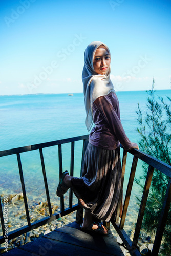 Cuadro en lienzo Full-length portrait of a stylish young Asian Muslim woman wearing a cream hijab, long-sleeved top, and pleated skirt posing playfully with one leg raised on a wooden deck
