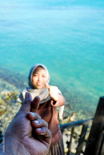 Canvas Print Close-up hand making a Korean finger heart sign in the foreground, with a young Asian Muslim woman in a hijab standing on a wooden ocean deck blurred in the background