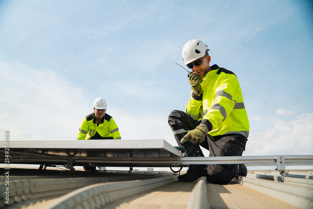 Fototapeta premium Solar panel technician worker roof safety helmet high visibility construction renewable energy installation maintenance on rooftop with two engineers using radio under blue sky