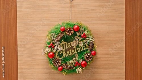 A man reaches out and adjusts a green Christmas wreath decorated with red baubles, pinecones and gold lettering hanging on a wooden door.