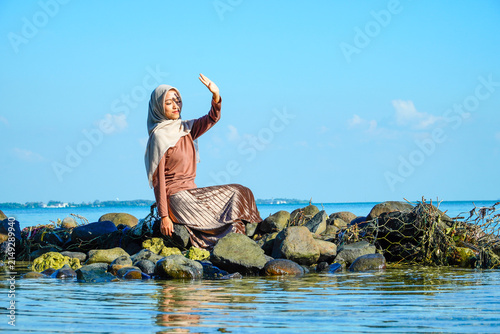Cuadro en lienzo ​A young Asian Muslim woman in a hijab and stylish pleated skirt sitting on coastal rocks, raising her hand to shield her eyes from intense sunlight