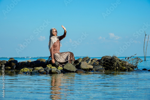 Картината върху платно ​A young Asian Muslim woman in a hijab and stylish pleated skirt sitting on coastal rocks, raising her hand to shield her eyes from intense sunlight