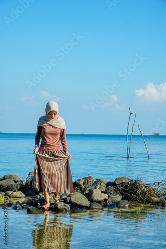 Photography ​A young Asian Muslim woman wearing a hijab and brown long-sleeved outfit carefully navigating mossy rocks along the shoreline