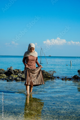 Fotografía ​A young Asian Muslim woman in a hijab holding her skirt while standing in clear, shallow ocean water near a rocky shore under a bright blue sky