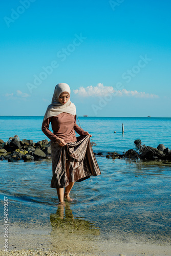 Quadro su tela ​A young Asian Muslim woman in a hijab holding her skirt while standing in clear, shallow ocean water near a rocky shore under a bright blue sky