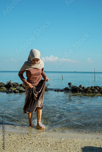 A young Asian Muslim woman in a hijab, long-sleeved shirt, and long skirt lifting her hem while walking barefoot through shallow, clear ocean water along a sandy beach under a bright blue sky. Concept