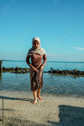 A young Asian Muslim woman in a hijab, long-sleeved shirt, and long skirt lifting her hem while walking barefoot through shallow, clear ocean water along a sandy beach under a bright blue sky. Concept