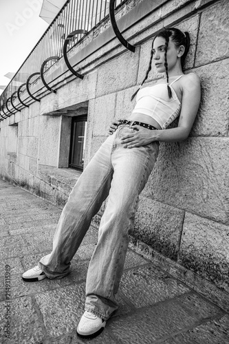 Low angle wide angle portrait of a trendy young woman in Y2K streetwear outfit with baggy jeans and crop top leaning against a stone building wall playing with her braided hair