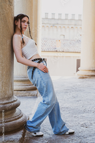 Monochrome black and white full body portrait of a trendy young woman with braided hair leaning against an ancient column looking in profile outdoors