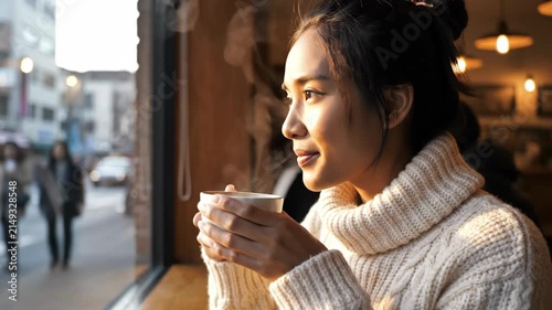 Young Asian woman in a white sweater sipping hot coffee by a cafe window during sunset.