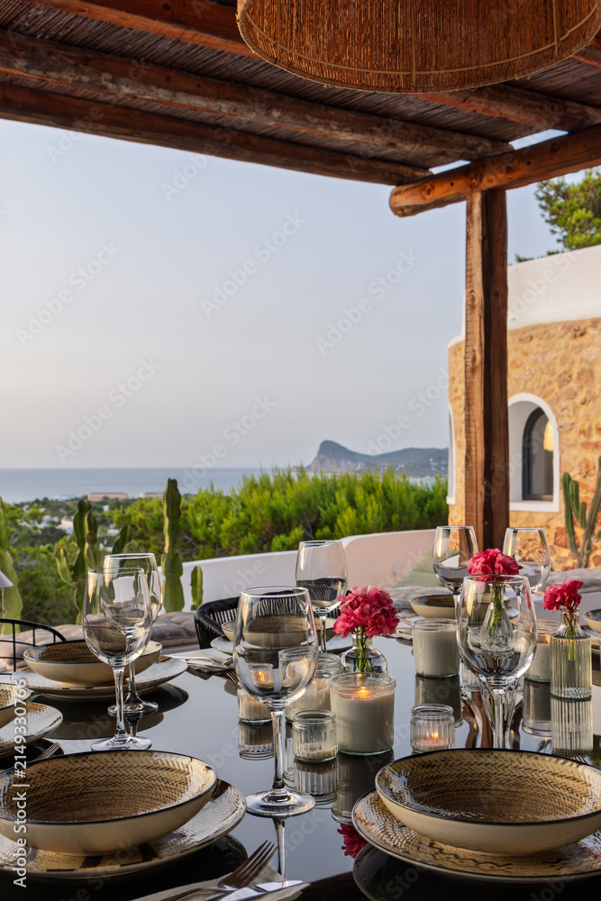 Fototapeta premium Candlelit dinner table sits in view on Ibiza, Spain with wine glasses, lit by warm twilight illumination and visible candle glow.
