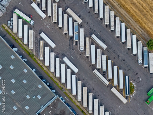 Aerial view of a logistics center in Sausheim, featuring rows of parked semi-trailer trucks and trailers in a paved depot area next to a warehouse. Sausheim, Grand Est, France.