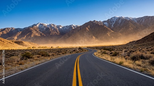 Scenic winding asphalt road through a desert valley toward majestic snow-capped mountains under a clear blue sky