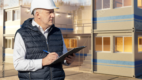 Modular Construction. Site engineer inspects modular building units and records project progress on a clipboard during residential development supervision.