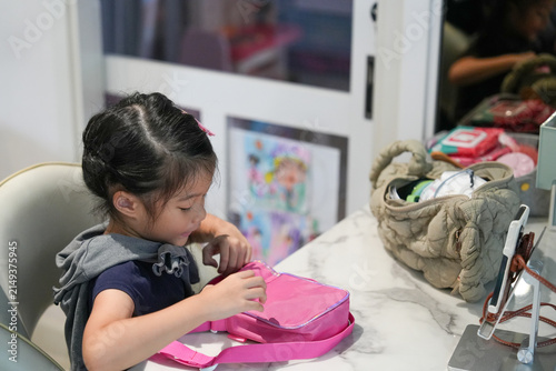 Little Asian Girl Opening Pink Bag at Desk Indoors,focused on opening a bright pink shoulder pouch or lunch bag while preparing or playing indoors.