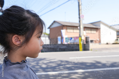 Side Profile of Little Asian Girl Looking Forward on Japanese Street, standing outdoors on a bright, sunny day with a Japanese neighborhood street and residential building in the background.