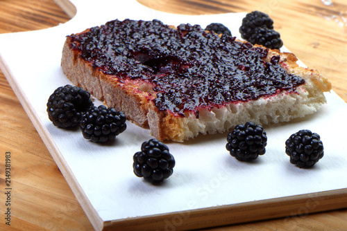 Close-up of a single slice of rustic bread with thick blackberry jam and fresh blackberries on a white wooden board