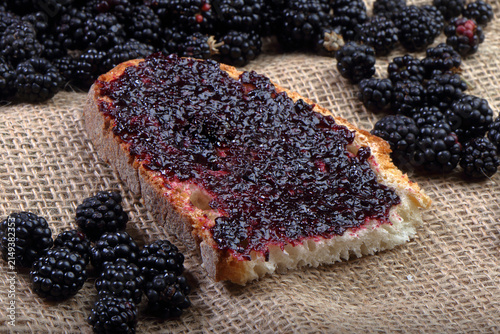 A slice of rustic bread spread with thick homemade blackberry jam surrounded by fresh blackberries on a burlap cloth