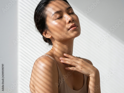 Beautiful young Asian woman with glowing skin and closed eyes enjoying sunlight with shadow patterns, skincare and natural beauty concept