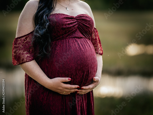 Pregnant woman in a burgundy lace maternity dress gently holding her baby bump during an outdoor maternity portrait session.