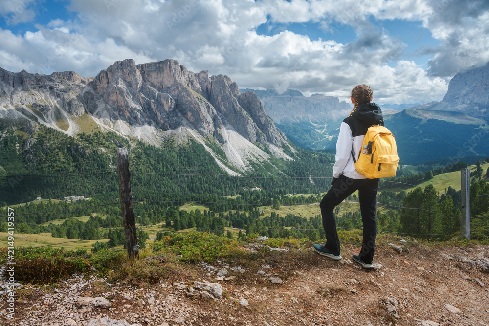 Obraz premium Woman hiker with yellow backpack standing on trail looking at mountain range in Dolomites, Italy. Alpine summer landscape with female tourist enjoying valley view, rocky peaks and cloudy sky. Hiking