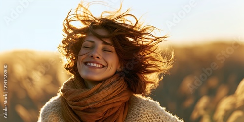 Portrait of a happy young woman with closed eyes smiling cheerfully while wind blows her long brown wavy hair outdoors in golden evening sunlight wearing warm scarf and sweater in nature field
