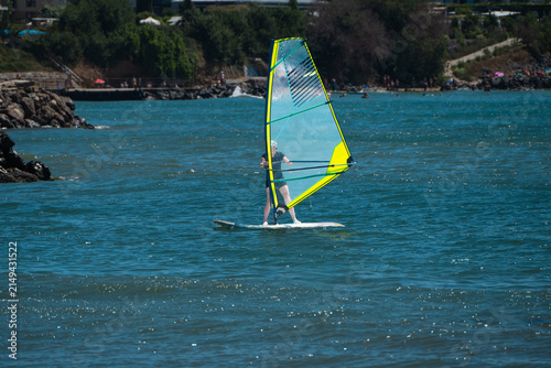 Foto A person enjoys a sunny summer day windsurfing on bright blue water with a yellow-edged transparent sail near a rocky, tree-lined coast