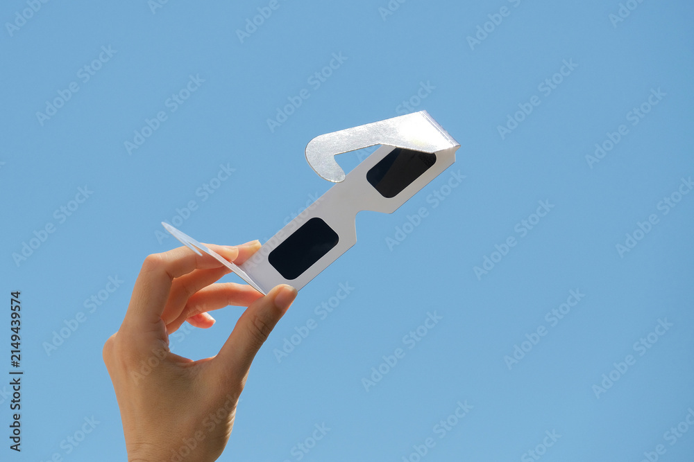 Obraz premium Woman hand holds solar eclipse glasses against blue sky background. Solar eclipse glasses are specially certified, ultra-dark glasses to protect eyes from damage when looking directly at sun.