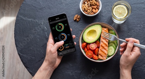 Keto diet tracking and healthy lifestyle concept with flat lay hands using smartphone app while eating salmon avocado bowl