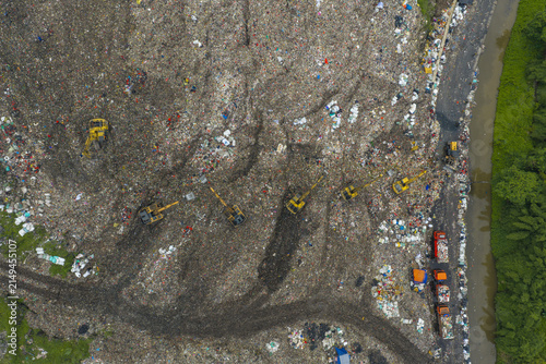 Aerial view of excavators working on a large landfill site with piles of waste and trucks parked along a river in Jakarta, Indonesia.