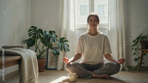 Young woman meditating in lotus pose on floor at home near window with house plants