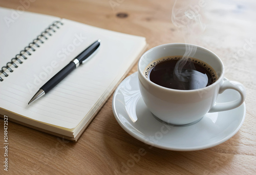 Steaming hot black coffee in a white cup with a pen and spiral notebook on a wooden desk, morning work