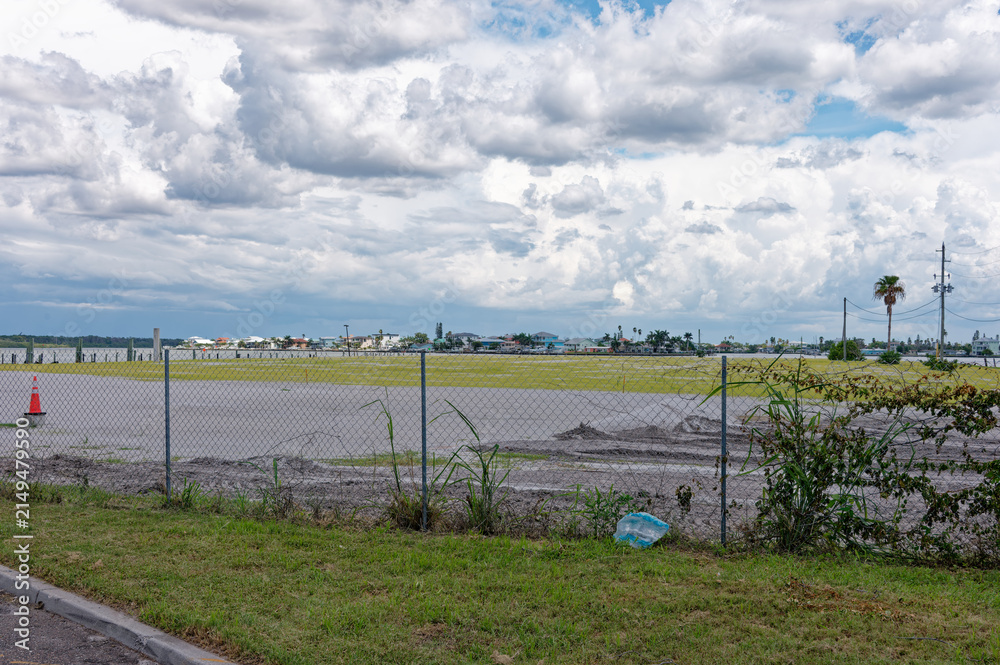 Fototapeta premium Chain link fence in the foreground frames an empty grassy field and coastal suburban homes in Madeira Beach, FL under a dramatic cloudy sky, conveying solitude, openness, and a moody late afternoon at