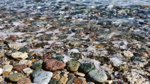 Gentle waves lap against shore of multi colored smooth river stones in cinematic high angle close up shot as clear water flows over pebbles on sunny day.