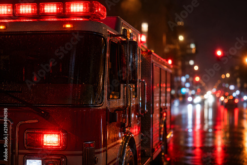Fire truck displays flashing red emergency lights on a wet city street at night, with traffic and rain reflections in the background.