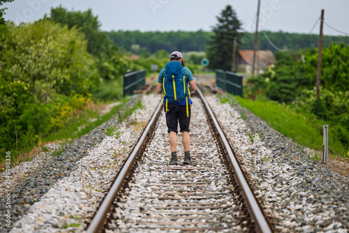 Solo Hiker With Blue Backpack Walking Along Rural Railway Tracks Through Green Countryside In Summer