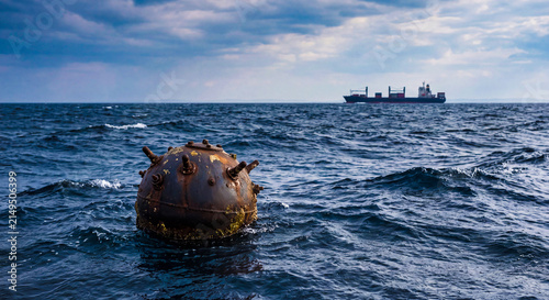 A contact naval mine floating on seawater. A spherical, rusty naval mine floating adrift and in the background a ship