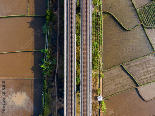 Aerial view of a double railway track cutting through lush green and brown rice paddies in the rural landscape of Banyumas Regency, Central Java, Indonesia.
