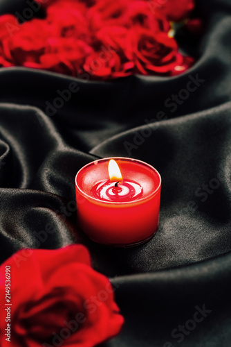 Obraz na płótnie Red candle with flame placed among red roses on dark fabric