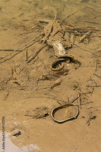 Foto Environmental pollution scene showing a discarded tin can, an old child shoe, and broken twigs in shallow, murky water on a sandy riverbed