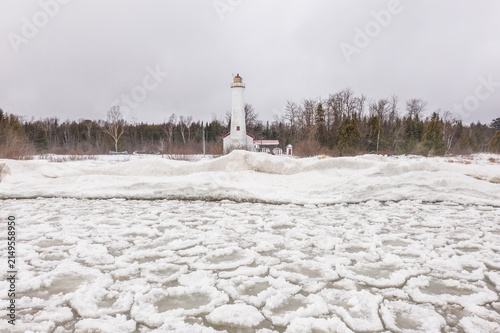 Aerial view of Sturgeon Point Lighthouse surrounded by ice-covered Lake Huron and snow-covered shoreline under a cloudy winter sky in Harrisville, Michigan, United States.