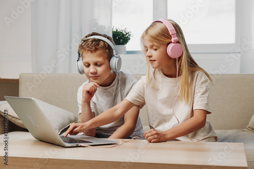Two children with headphones using laptop on table in cozy living room. Concept of technology, education, and entertainment at home for kids.