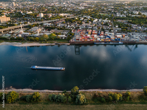 Aerial view of Hafen Bonn featuring a cargo barge navigating the Rhine river alongside industrial harbor facilities with shipping containers in Bonn, Nordrhein-Westfalen, Germany.