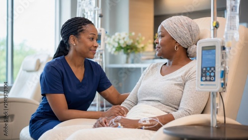 Female oncology nurse in blue scrubs provides emotional support to a cancer patient wearing a headscarf during treatment in a bright, modern infusion room