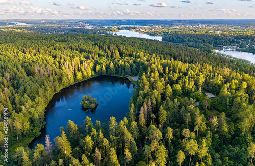 Aerial drone photo of a small pond in forest with a small island in Finland Hämeenlinna Aulanko Joutsenlampi with city in distant background. 