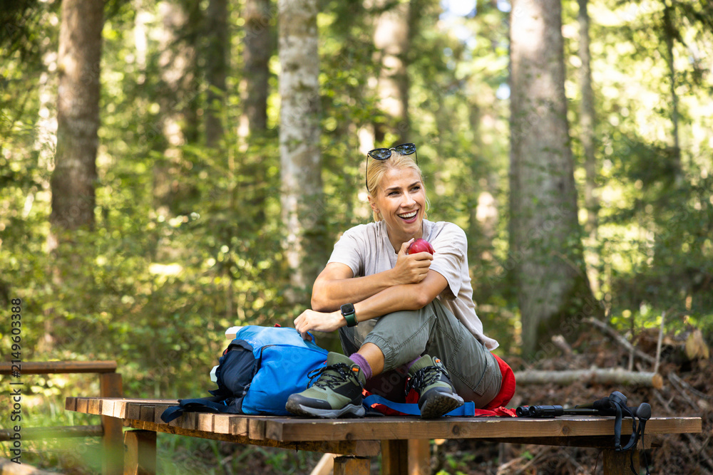 Fototapeta premium Woman hiker relaxing on a wooden bench, eating a fresh apple and smiling, taking a break in a green forest