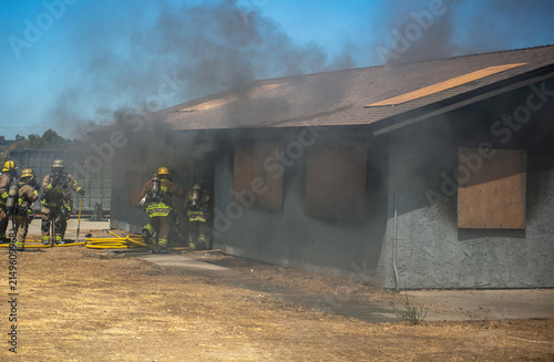 Firemen and women practicing to put out fires on an abandoned house looking at fire fighters preparing to enter a practice building with their equipment to rescue people inside and put out the fire.