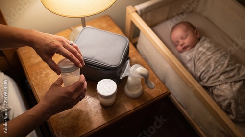 Mother prepares milk bottle next to sleeping baby and breast pump at night