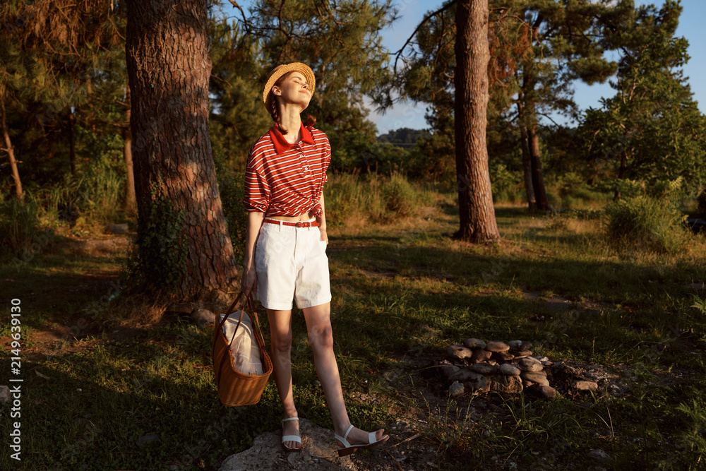Obraz premium A redheaded woman lifestyle enjoying golden hour in a forest park. Wearing straw hat and summer clothes holding a picnic basket. Concept for outdoor leisure, relaxation, and nature travel.