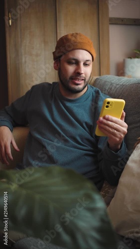 Young Latino man sitting on his sofa while making a video call on his smartphone, representing digital communication, social connection, mobile technology, and everyday life at home.
